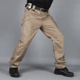 Hombre con polera negra, cinturón con placa metálica en la cadera y botas negras, en posición abierta sobre fondo gris.