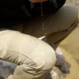 Rodilla de una persona en cuclillas con pantalón beige, con gotas de agua cayendo y quedando sobre la tela.