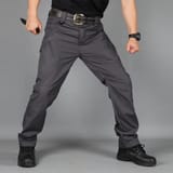 Hombre con polera negra, cinturón de hebilla dorada y botas negras, en posición abierta sobre fondo gris, sosteniendo un cuchillo negro.