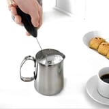 Mano sostiene el espumador negro dentro de un jarro de acero con leche, sobre mesa blanca con galletas y taza de café.