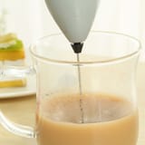 Espumador plateado metido en una taza de vidrio con café con leche, formando espuma en la superficie.