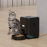 Gato sentado en el piso junto a un comedero automático negro conectado con cable y baterías.