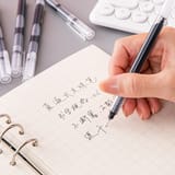 Mano escribiendo con un bolígrafo en un cuaderno de cuadrícula junto a un teclado.