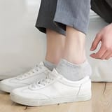 Pies con calcetines tobilleros grises puestos con zapatillas blancas, piernas con pantalón gris, una mano cerca del tobillo.