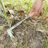 Una mano empuña una herramienta de hoja plana con mango de madera, hundida en la tierra suelta entre pasto seco.