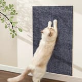 Panel rectangular de alfombra oscura pegado en una muralla blanca, con un gato estirándose para arañarlo y sombras de hojas al lado.