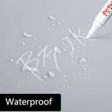 Punta de marcador blanco escribiendo letras sobre una superficie gris con gotas de agua.